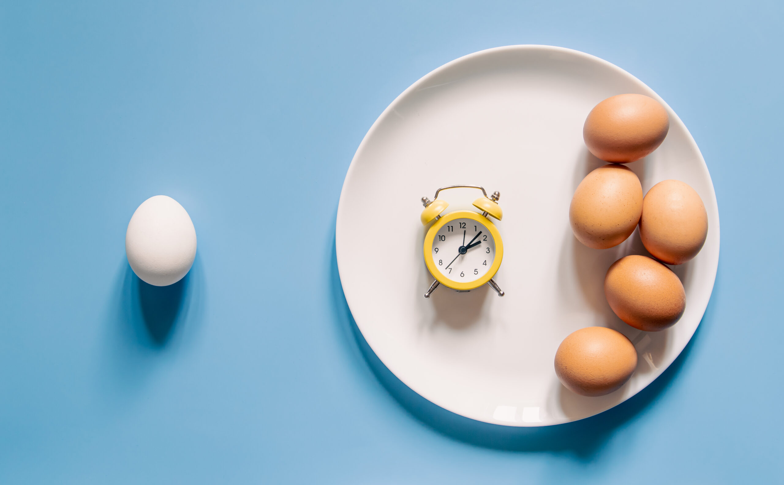 Image Plate with eggs and an alarm clock on a blue background, flat lay.